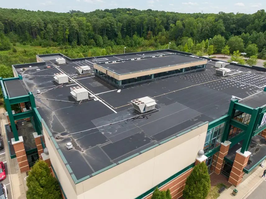Aerial view of a large, rectangular building with a flat roof. The roof has multiple HVAC units and piping. The structure is surrounded by trees and greenery, indicating a location near a wooded area.