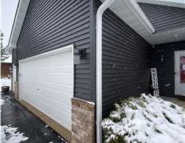 close up of gray siding on a garage, snow is falling and collecting on the bushes around the garage
