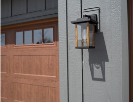 A moder exterior light fixture that is installed on the side of a garage on a gray house. The background is another garage that is colored brown.