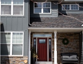 A red front door is the center of this image, to the right is a stone wall, the rest of the two story home is made up of gray siding and big windows. A comfy outdoor couch sits on the front porch.