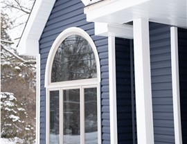 close up of large windows, half circle window and three rectangle panes of glass, around this window is dark blue vinyl siding. Behind this portion of the house are snowy trees