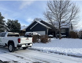 gray home with roof and yard covered in snow, there is a car parked int eh driveway also covered in snow and a white truck parked outside