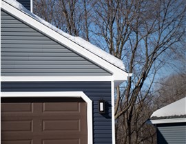 the left half of this image is a close up of a garage with brown doors, around the doors is dark blue siding and the elevation above the garage is gray. Behind the garage are bare trees