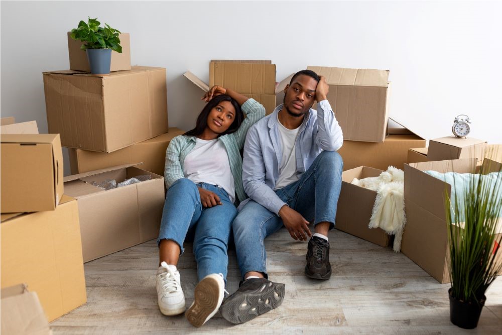 Two people sit on the floor surrounded by moving boxes