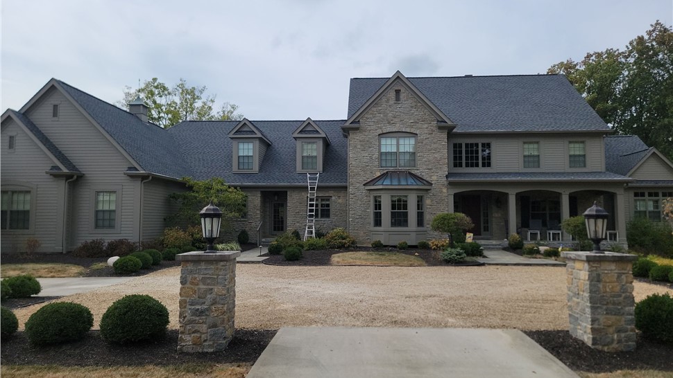 A large two-story home in Indian Hill, OH, with a newly installed Charcoal Timberline UHDZ shingle roof from GAF. The house features a mix of stone and siding, dormer windows, a covered front porch, and a well-maintained landscape with stone pillars at the entrance. A ladder is visible, suggesting recent roofing work.