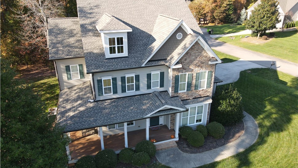 Aerial view of a Wake Forest, NC, home with a newly installed Timberline HDZ roof, featuring beige siding, stone accents, green shutters, a covered porch, and a curved driveway, surrounded by a green lawn and trees.