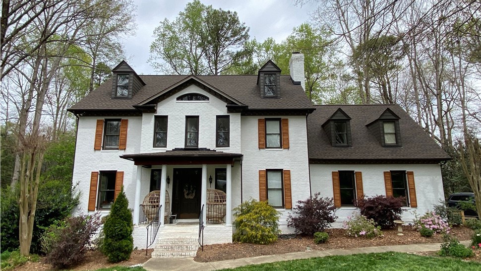 A two-story white brick home in Holly Springs, NC, featuring a newly installed Barkwood Timberline HDZ shingle roof, wooden shutters, dormer windows, a welcoming front porch, and a beautifully landscaped lawn surrounded by tall trees.