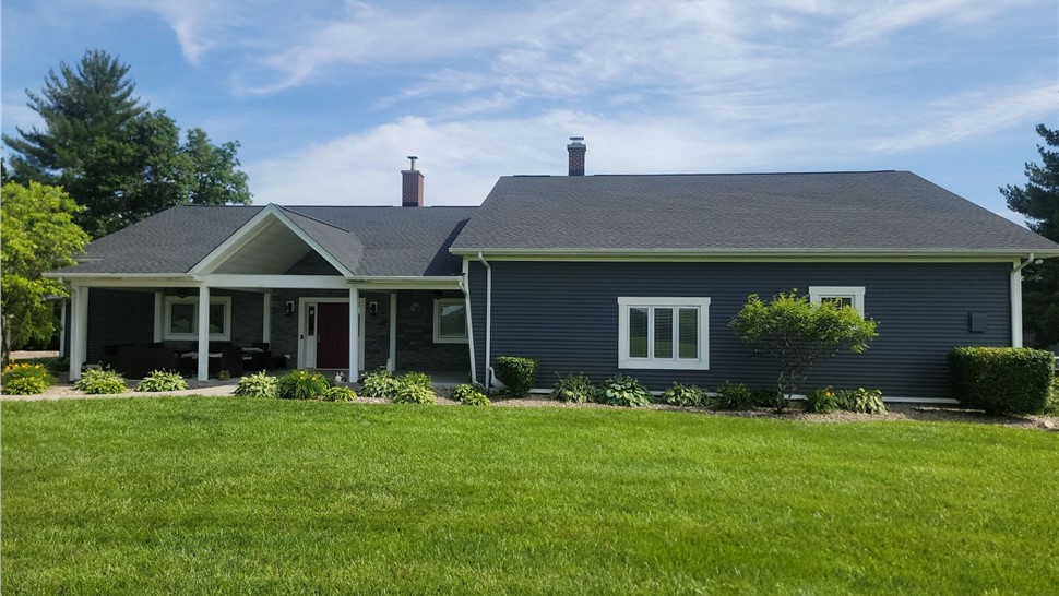 A home in Hamilton, OH, with a new Charcoal Timberline HDZ shingle roof. The house features navy blue siding, white trim, a covered porch, and a well-maintained green lawn under a bright blue sky.