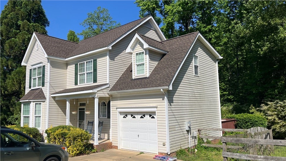 A two-story beige home in Apex, NC, with a newly installed Barkwood Timberline HDZ shingle roof. The house features white trim, green shutters, a covered front porch with rocking chairs, a single-car garage, and a backdrop of lush greenery and tall trees under a clear blue sky.