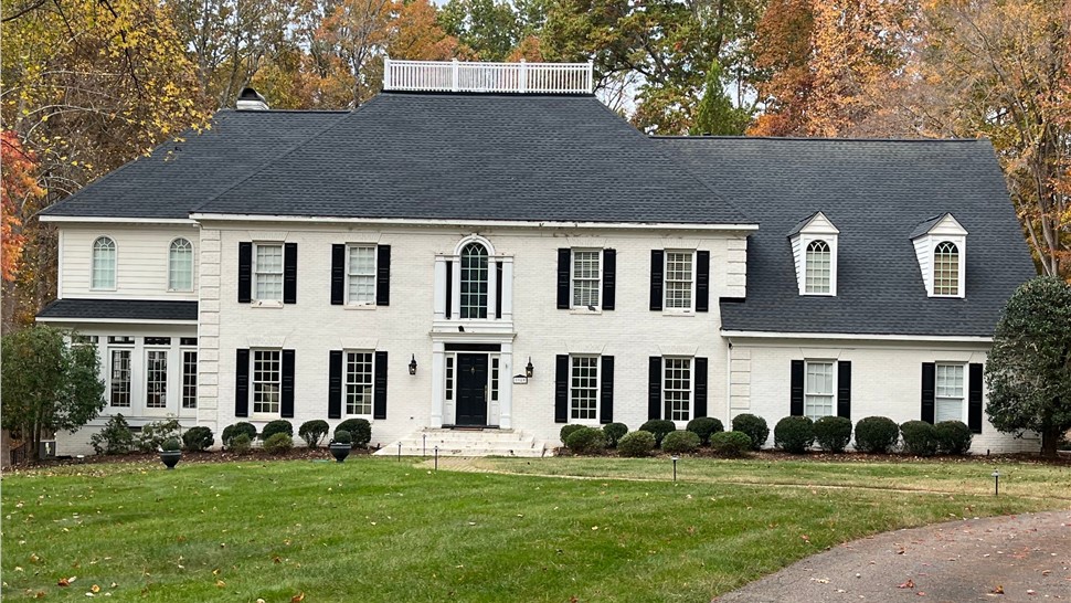 Colonial-style home in Raleigh, NC, with a newly installed Charcoal Timberline HDZ shingle roof, white brick exterior, black shutters, a rooftop deck, and a well-maintained landscape with a curved driveway and autumn trees.