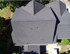 Aerial view of a residential home in Holly Springs, NC, featuring a newly installed Charcoal Timberline HDZ shingle roof with a multi-sectioned design, surrounded by lush greenery, a backyard patio, and a driveway.