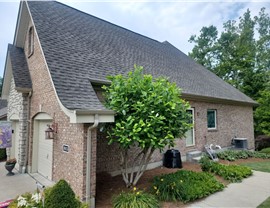 A Cleves, Ohio, home with a newly installed roof featuring Weathered Wood Timberline HDZ shingles, complemented by a brick exterior and vibrant landscaping with shrubs, flowers, and a small tree.