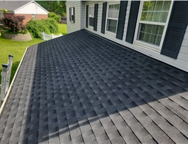 Close-up view of a newly installed roof with Charcoal Timberline HDZ shingles on a light-colored house with black shutters in Fairfield, OH. The roof has a clean and polished finish, with a lush green yard visible in the background.