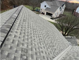 A new roof installation in Milford, OH, featuring Weathered Wood Timberline HDZ shingles. The image shows clean shingle alignment and ridge caps with a suburban home and wooded background under a cloudy sky.