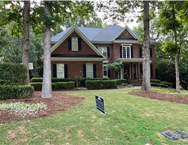 A two-story red brick home in Cary, NC, with a newly installed Charcoal Timberline HDZ shingle roof. The house features black shutters, a covered front porch with white columns, lush landscaping, and tall trees. A Feazel Roofing sign is placed in the front yard.
