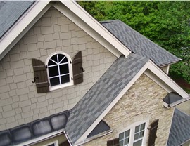 A close-up view of a Holly Springs, NC, home with Nantucket Morning Timberline HDZ shingles, stone and shingle siding, an arched window with shutters, and white trim, surrounded by lush greenery.