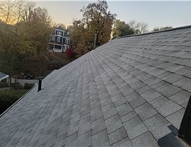 Close-up view of a Hyde Park, OH, roof replacement using Pewter Grey Timberline HDZ shingles, with a classic blue house and autumn trees in the background at dusk.