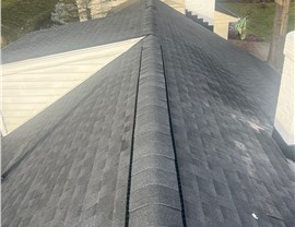 Close-up of a Charcoal Timberline HDZ shingle roof replacement in Hyde Park, OH, highlighting the ridge vent and neatly installed shingles. The background features a light-colored house, chimney, and trees.