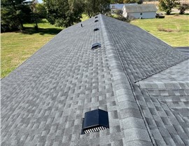 A newly installed Charcoal Timberline HDZ shingle roof in Hamilton, Ohio, with black roof vents evenly spaced along the ridge. The background features a green yard, trees, and a clear blue sky with scattered clouds.