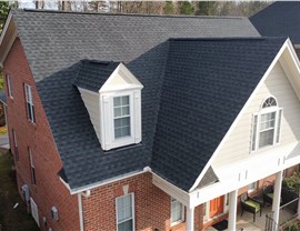 Aerial view of a brick home in Apex, NC, featuring a newly installed Charcoal Timberline HDZ shingle roof. The dark shingles contrast with the red brick and white trim. The roof has multiple gables, a dormer window, and a covered front porch with white columns and black patio furniture. Lush greenery surrounds the home.