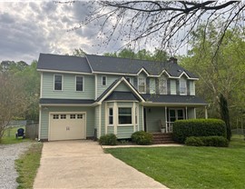 A two-story green home in Cary, NC, with a newly installed Charcoal Timberline HDZ shingle roof. The dark shingles contrast with the green siding and white trim. The house features a front porch with rocking chairs, a bay window, an attached garage, and a well-maintained lawn with trees and hedges. The sky is partly cloudy.