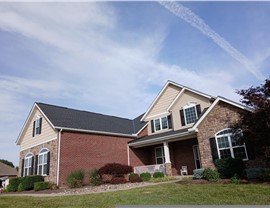 A Hamilton, Ohio home with a new Charcoal Timberline HDZ shingle roof, featuring a mix of brick, siding, and stone accents. The front yard is landscaped with greenery and flowers, under a bright blue sky with wispy clouds.