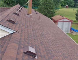 Newly installed Hickory Timberline HDZ shingle roof in Goshen, Ohio, featuring box vents and a chimney pipe. The reddish-brown shingles contrast with the surrounding greenery, with a shed and open backyard visible in the background.