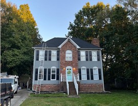A two-story brick and blue-gray siding home in Kernersville, NC, with a newly installed Charcoal Timberline HDZ shingle roof. The house features black shutters, a turquoise front door, and a white-railed entrance staircase, set against a background of a partly cloudy sky.