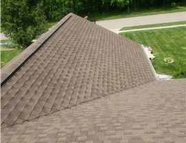 New Hickory Timberline HDZ shingle roof in Goshen, Ohio, featuring a ridge vent for ventilation. The brown-toned shingles contrast with the green surroundings. A residential street and lawn are visible in the background, along with roofing materials near the edge.