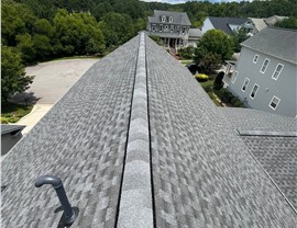 Close-up view of a newly installed Pewter Gray Timberline HDZ roof in Wake Forest, NC, showing the ridge vent, architectural shingles, and surrounding neighborhood with greenery and homes under a partly cloudy sky.