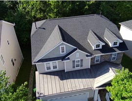 Aerial view of a Raleigh, NC, home with newly installed Charcoal Timberline HDZ shingles, light gray siding, stone accents, white trim, and a metal porch roof, surrounded by trees and neighboring houses.