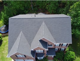 Aerial view of a Cary, NC brick home with a new Charcoal Timberline HDZ shingle roof, featuring multiple gables, black accent roofing, and a well-maintained backyard with trees and a trampoline.