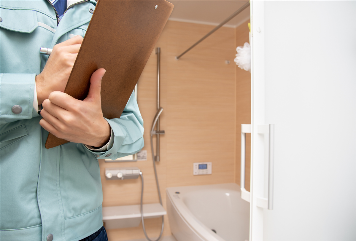A contractor in a light blue uniform holding a clipboard while inspecting a bathroom with a white soaking tub, handheld showerhead, and wood-look wall panels