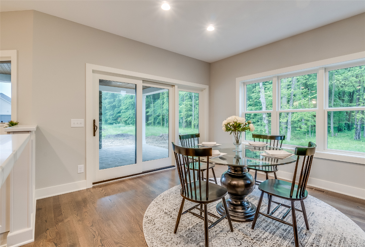 A bright dining area with hardwood floors featuring a round glass-top table with black Windsor chairs on a circular rug, surrounded by large sliding glass doors and windows overlooking a wooded backyard.