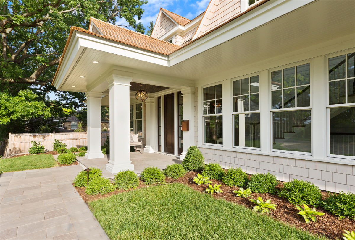 Exterior front entrance of a modern shingle-style home featuring a covered porch with white square columns, a dark wood front door, a brass globe pendant light, and a cedar shake roof. The landscaping includes neatly trimmed boxwood shrubs, hostas, and a stone paver walkway bordered by a green lawn and mature trees