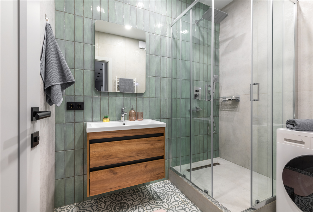 Modern compact bathroom with glossy sage green square wall tiles, a floating wood vanity with black hardware, a rectangular mirror, glass-enclosed walk-in shower with a rain showerhead, patterned encaustic floor tiles, and a washer-dryer unit tucked beside the shower