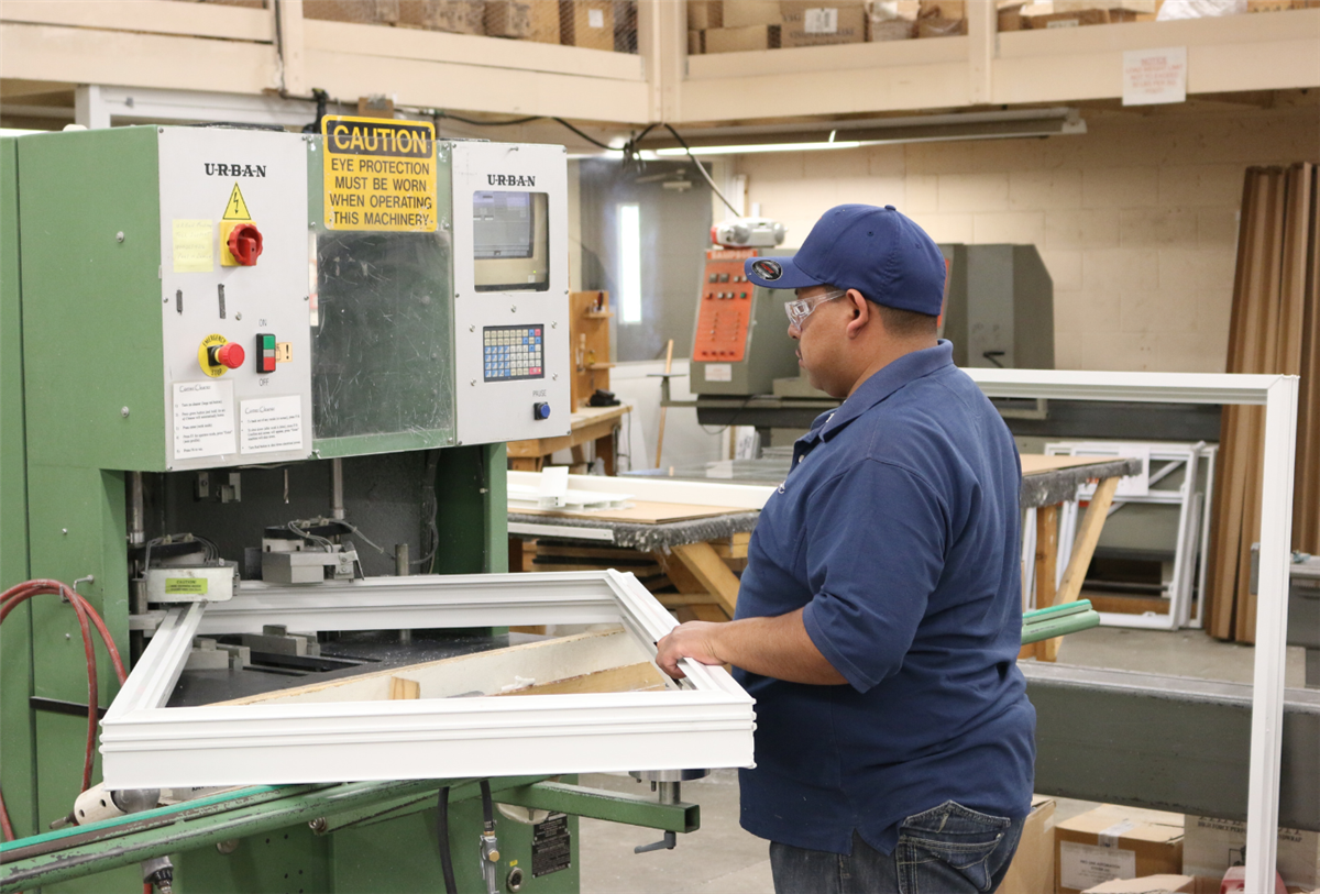A factory worker wearing safety glasses operating an Urban window manufacturing machine while assembling a white vinyl window frame in a production facility