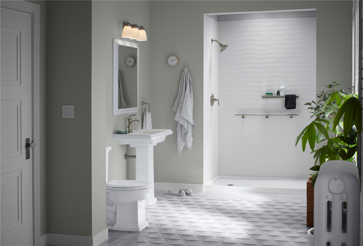 Spacious sage green bathroom featuring a Reveal by KOHLER walk-in shower with White Brick LuxStone walls, a pedestal sink with vanity mirror, toilet, geometric grey floor tile, and large indoor plants