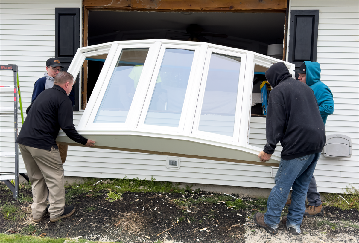 A four-person crew carefully lifting and positioning a large white bow window unit into a rough opening on the exterior of a white vinyl-sided home