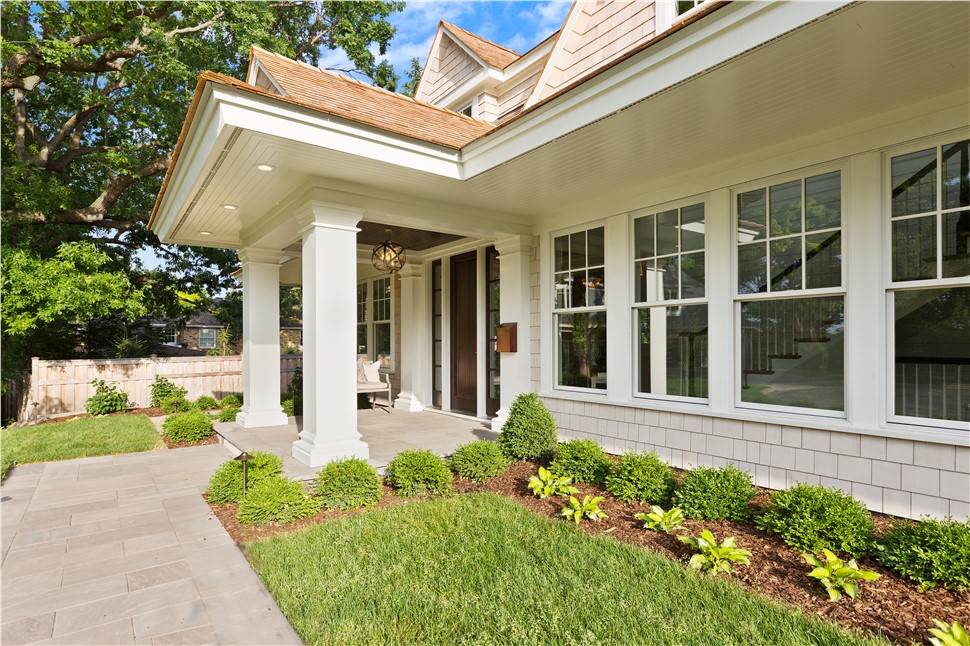 Exterior front entrance of a modern shingle-style home featuring a covered porch with white square columns, a dark wood front door, a brass globe pendant light, and a cedar shake roof. The landscaping includes neatly trimmed boxwood shrubs, hostas, and a stone paver walkway bordered by a green lawn and mature trees