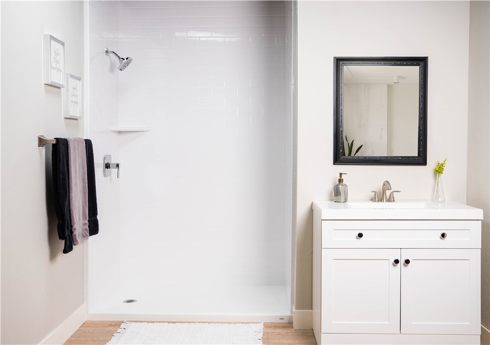 A clean white bathroom featuring an open walk-in shower with white subway tile walls and a chrome showerhead, a white shaker-style vanity with brushed nickel faucet, a black framed mirror, folded towels on a towel bar, and wood-look flooring