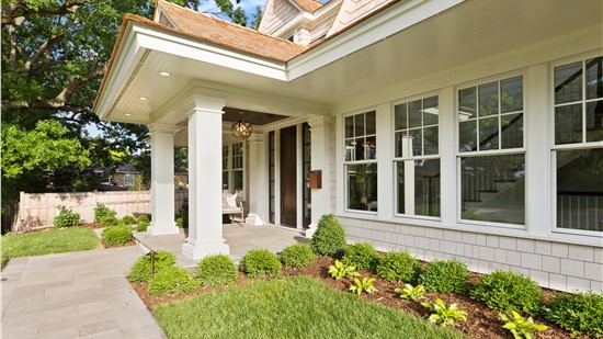 Exterior front entrance of a modern shingle-style home featuring a covered porch with white square columns, a dark wood front door, a brass globe pendant light, and a cedar shake roof. The landscaping includes neatly trimmed boxwood shrubs, hostas, and a stone paver walkway bordered by a green lawn and mature trees