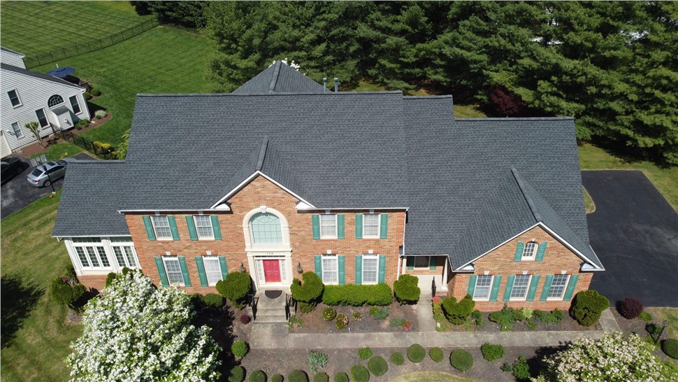 Aerial view of a brick home with a new roof featuring Charcoal Timberline HDZ shingles, surrounded by green lawns, a blooming tree, and a paved driveway.