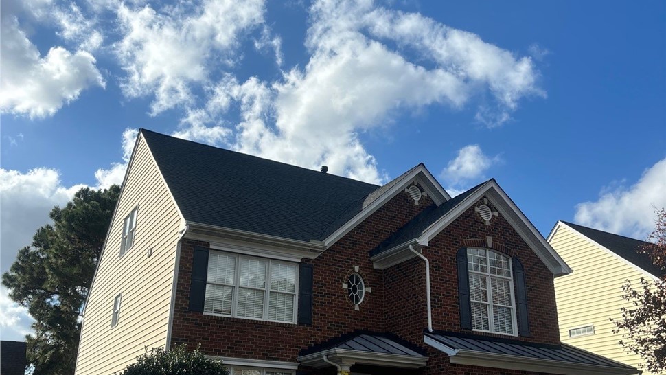 Front view of a two-story brick home with a new Charcoal Timberline HDZ roof, white trim, black shutters, and a decorated lawn with Halloween-themed ornaments. The house is bordered by a hedge-lined driveway, under a blue sky with scattered clouds in Glen Allen, VA.