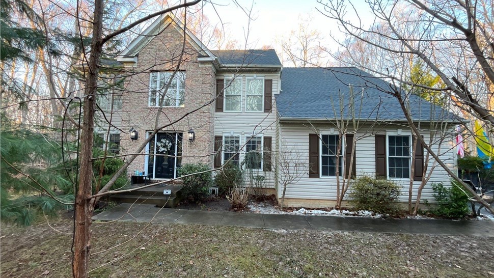 Two-story home in Manassas, VA, with a new Charcoal Timberline HDZ shingle roof. The house features a brick facade, cream-colored siding, brown shutters, and a front pathway surrounded by bare trees and shrubs during winter.