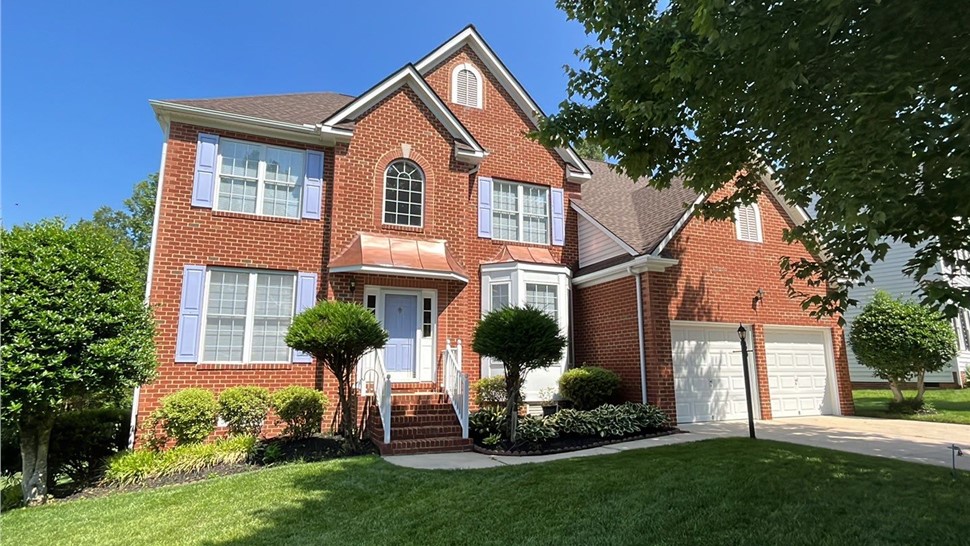 Elegant brick home in Chesterfield, VA, with a new roof made of Barkwood Timberline HDZ shingles, complemented by lavender shutters, a copper accent above the entrance, and lush landscaping under a clear blue sky.