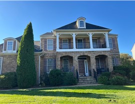 Front view of a two-story stone-front house with a new Charcoal Timberline HDZ roof, a covered balcony with white columns and black railings, brown shutters, and a solid wood front door. The home is surrounded by a neatly manicured lawn, a tall evergreen, and vibrant shrubs under a bright blue sky in Glen Allen, VA.