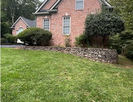 Brick home in Manassas, Virginia, with a freshly installed Charcoal Timberline HDZ shingle roof. The house has a large, green lawn, trimmed bushes, and a stacked stone retaining wall, creating a timeless and well-maintained look.