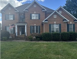 Front view of a two-story brick home with a new Weathered Wood Timberline HDZ roof, green shutters, and white trim. The entryway includes a white-columned portico, and an American flag is displayed near the front door. The lawn is bordered by neatly trimmed hedges, and the sky is blue with scattered clouds in Glen Allen, VA.