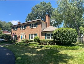 Two-story brick home in Alexandria, VA, with a new roof featuring Weathered Wood Timberline HDZ shingles that complement the brick's color tone. The house includes green shutters, white-trimmed windows, and lush landscaping in the front yard.